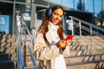 Optimistic woman wearing headphones and holding phone outdoors. Young woman listens to music, audiobook or podcast while walking on sunny street. Communication in social networks. Lifestyle.