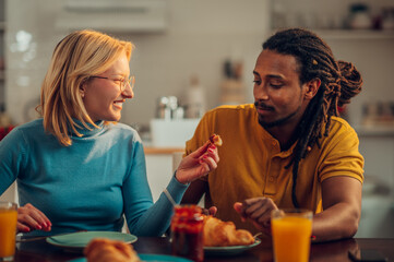 A happy interracial couple is sitting at the dining table at home and having croissants for breakfast.