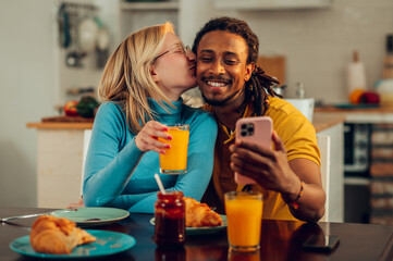 A young woman is kissing her multiracial husband while he is taking selfies at the breakfast table.