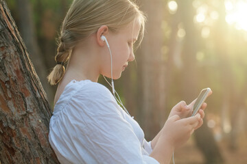 Teenage girl in white shirt and headphones typing on mobile phone and standing sideways near tree on the background of forest and  sunlight