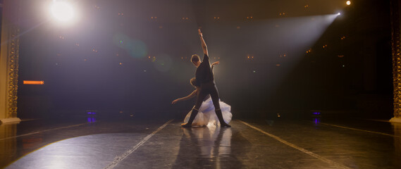 Static full shot of ballet dancers practicing graceful ballet movements and rehearse choreography on classic theater stage illuminated by spotlights. Dance partners prepare theatrical performance.
