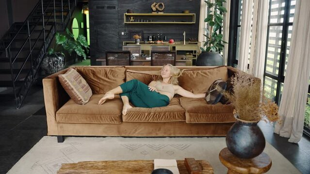 Concept for real estate sales, tenancy rental business, happy customer bought new house apartment or villa. Young blond woman enjoys new home, sitting on sofa in big living room with designed interior