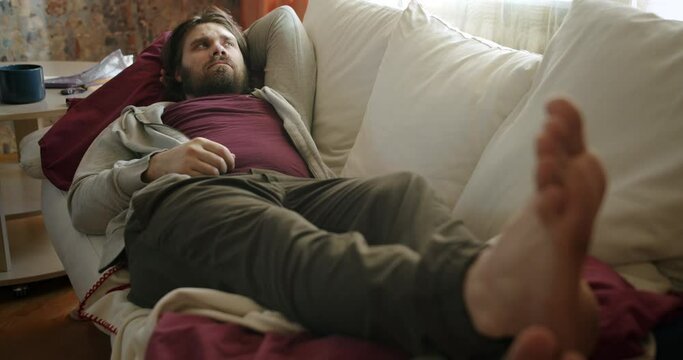 Carefree, careless lazy beard man on the couch staring at the ceiling, looking around out of boredom and idleness. Slacker thinking about procrastination and wasting time. Boredom, laziness concept