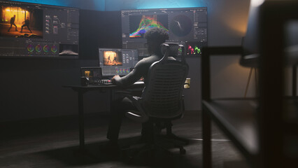 Handheld shot of African American colorist doing color grading on computer in modern studio. Multiple big monitors with action movie footage and program interface on the wall. Video post production.