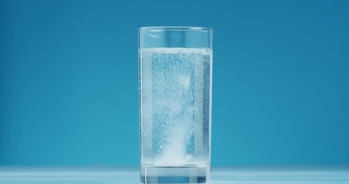 Glass of water falling white round tablet of soluble aspirin bubbling in glass of water on blue background. Effervescent tablet of acetylsalicylic acid dissolves in water forming many hiss bubbles.
