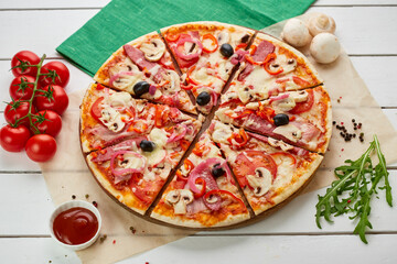Freshly baked pizza with smoked sausages, red pepper, mushrooms, onion and olives served on wooden background with tomatoes, sauce and herbs. Food delivery concept. Restaurant menu