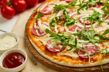 Freshly baked tasty pepperoni pizza with salami, mozzarella cheese and rukkola served on wooden background with tomatoes, sauce and herbs. Food delivery concept. Restaurant menu