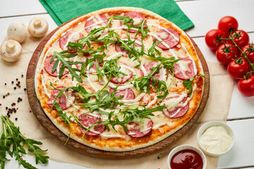 Freshly baked tasty pepperoni pizza with salami, mozzarella cheese and rukkola served on wooden background with tomatoes, sauce and herbs. Food delivery concept. Restaurant menu