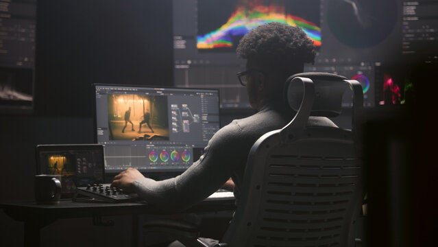 African American film editor does color grading on computer in modern studio. Big monitors with RGB colour correction graphic bar and program interface on the wall. Video post production. Handheld.