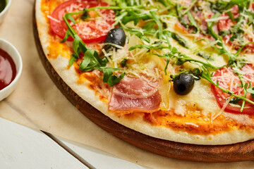 Freshly baked pizza with ham, rukkola, sauce pesto and olives served on wooden background with tomatoes, sauces and herbs. Food delivery concept. Restaurant menu