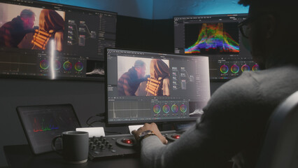 African American movie maker does color grading on computer in studio with equipment. Big monitors with action film footage and professional software interface on wall. Video post production process.