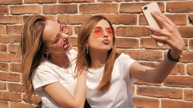 Two young smiling hipster blond women in summer white t-shirt clothes. Girls taking selfie self portrait photos on smartphone. Models posing on street background. Female showing positive face emotions