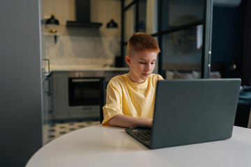Focused redhead 10 years old boy with freckles looking to laptop screen and typing doing homework sitting at table on background of kitchen. Cute teen boy surfing internet on notebook computer at home