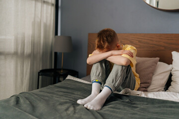 Sad unrecognizable little boy hugging knees, sobbing with head bowed and crying sitting on bed in children room. Stressed child feeling pressure, thinking of kid problems. Concept of child depression.