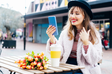 Stylish young smiling woman enjoying coffee from a reusable cup and having video call on her phone in a street cafe. Fresh tulips bouquet on the table. Springtime positive mood, street fashion.