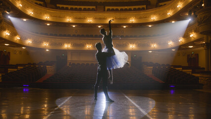 Couple of classical ballet dancers practice on theatre stage before performance. Man in training suit lifts graceful ballerina. Dance choreography rehearsal. Illuminated theatrical hall. Slow motion.