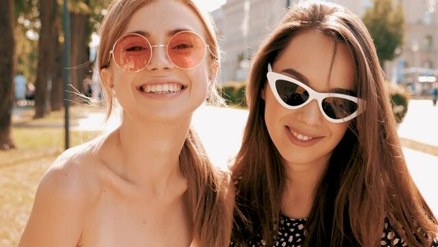 Portrait of two young beautiful smiling hipster girls in trendy summer clothes.Carefree women posing on street background. Positive models having fun in sunglasses and blowing bubble with chewing gum