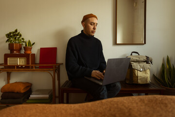 A caucasian man sitting on a bench in a bedroom writing on a laptop.