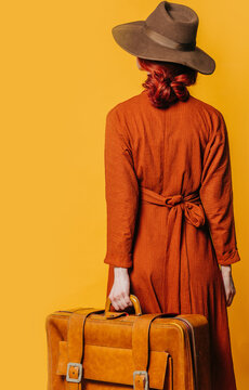 Back side view on stylish redheaded woman in hat and brown dress with suitcase on yellow background