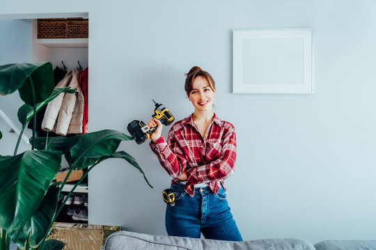 Happy Smiling Proud Of Herself Young Brunette Woman In Plaid Shirt Holding Electrical Screwdriver After She Put A Picture On The Wall At Her Home. Housekeeping Work. Doing Repair Herself. DIY Concept.