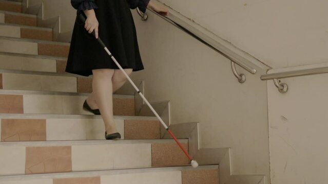 Woman with blindness disability walking down stairs using blind walking stick or long cane and touching stair handrail