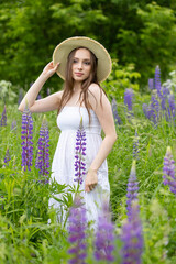 Beautiful young girl in a white dress, straw hat with a bouquet of violet flowers in her hands and picnic basket. Pretty woman in summer in the blooming field holding a bunch of purple lupin