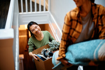 Happy Asian woman and her friend carrying their belongings upstairs while relocating into new...