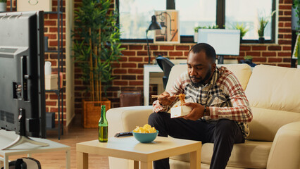 Smiling person eating noodles with chopsticks on sofa, enjoying asian meal from delivery. Male adult relaxing with bottle of alcohol and takeaway food, laughing at favorite movie. Tripod shot.