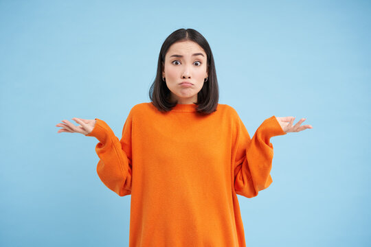 I dont know. Puzzled asian woman shrugs shoulders with hands spread sideways, confused, dont know what to do, clueless and unaware what happened, blue background