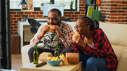 Modern young couple eating burgers together on couch, enjoying hamburgers with fries and bottles of alcohol. Cheerful man and woman having fun watching film on television, drinking beer.