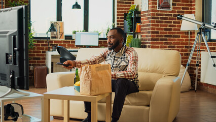 African american man opening food delivery bag in living room, preparing to eat takeout fast food in front of television. Male adult having takeaway dinner watching film on tv at home.