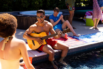 Happy diverse group of friends having pool party, playing guitar in garden