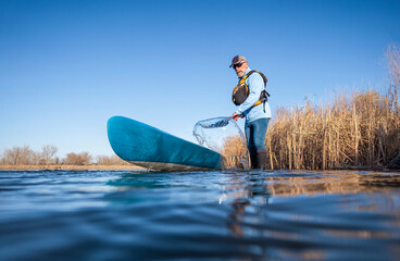 senior male paddler is rinsing his  stand up paddleboard after paddling on on a calm lake in early...