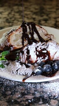 pouring chocolate on a croissant already sprinkled with powdered sugar with blueberries or lokhina mint leaves a lot of sweets around the plate powdered sugar is also scattered on the marble table