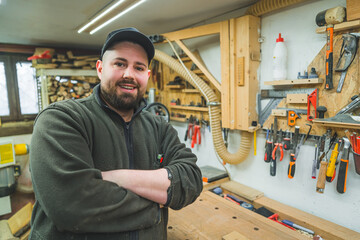 Positive male carpenter in woodworking garage standing with folded arms and smiling at camera. Variety of tools used in work with wood. High quality photo