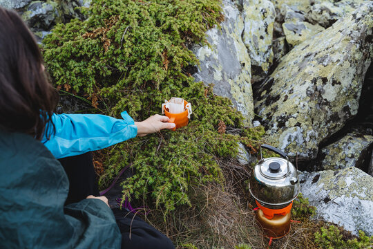 A girl on a hike brews coffee in a filter bag, a kettle stands on a burner, a tourist prepares breakfast at a campsite, a mountain hike alone, a hot drink.