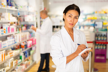Female Asian pharmacist in white gown standing in salesroom of chemist shop with pen and sheets of paper in hands and looking in camera.