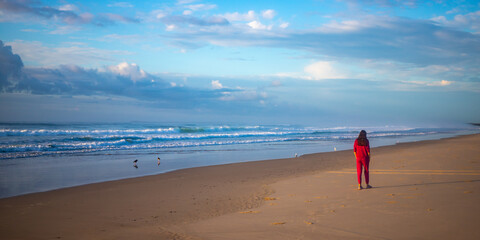 Girl in pink tracksuit walks on empty Ten Mile Beach during late surise. Black rocks campsite,...