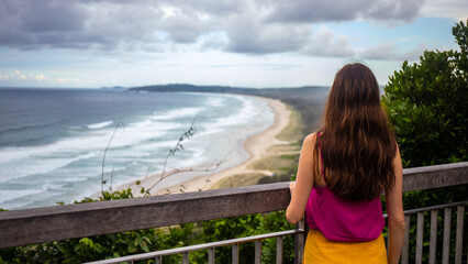 Fototapeta na wymiar Beautiful long-haired girl stands admiring the magnificent rocky coastline and beaches in the famous Byron Bay, New South Wales, Australia