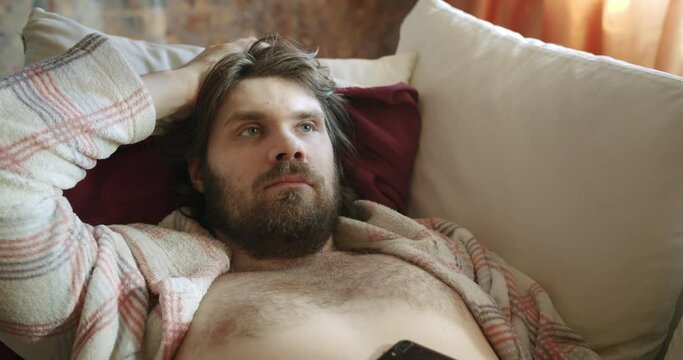 Carefree, careless lazy beard man in robe lying on the couch scratching head. Unkept, shabby, pensive look. Procrastination, waste time and motivation problem. Laziness and procrastination concept