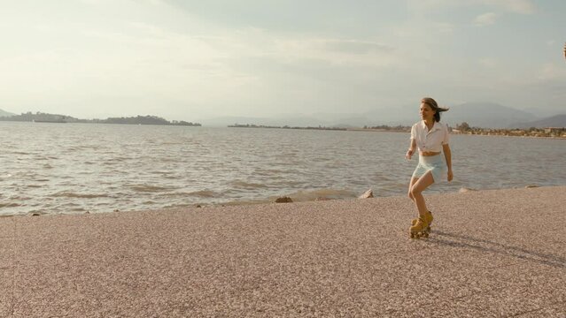 Cheerful carefree young woman rollerblading along coastline. Slim athletic woman roller-skate on retro quads roller skates near sea
