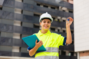 Young woman engineer in hard hat on building site showing key from new home