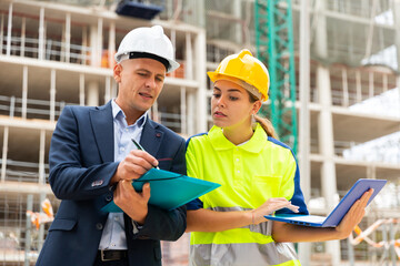 Engineer man in suit and hardhat discussing with female worker project documentation while they're...