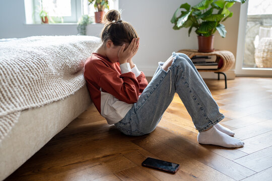 Unhappy teen girl covering face with hands and crying while sitting on floor with mobile phone nearby, upset frustrated child teenager being bullied or harassed online. Cyberbullying among teens