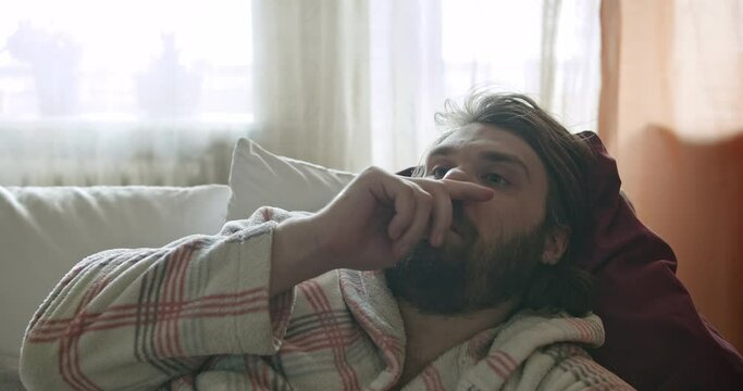 Carefree, careless lazy beard man lying on the couch. Unkept, compulsive nose picking. Thinking about procrastination, waste time and motivation. Laziness and procrastination concept