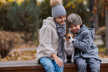 Brother and sister sit in an embrace with a dog on a bench for a walk in the autumn park. Boy, girl and jack russell terrier. 