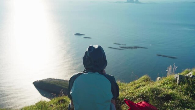 Rear Of A Person In A Hoody Sitting On A The Mountain Edge In Lovund Island, Norway. High Angle