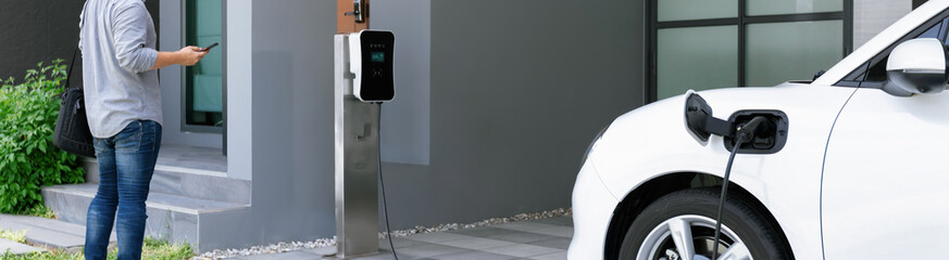 Progressive asian man and electric car with home charging station. Concept of the use of electric...