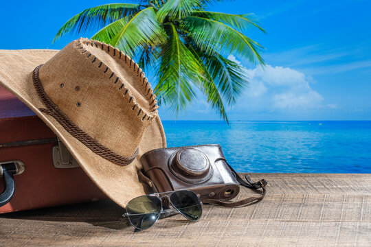 Suitcase, sun hat, photo camera and sunglasses with sea water, coconut palm tree and blue sky background on sunny summer day in tropical beach, copy space. Vacation and travel concept