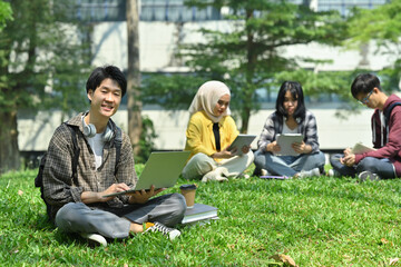 Fototapeta premium Young asian man student using laptop on green grass in front of university building. Education, technology and lifestyle concept